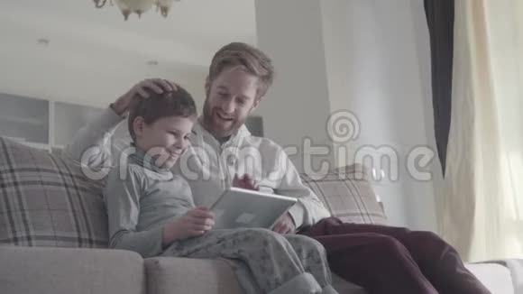 微笑快乐的父子用数码平板电脑坐在大客厅的沙发上快乐的家庭花时间视频的预览图