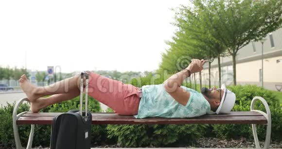 从侧面可以看到一个为度假做好准备的迷人的非洲男人躺在长凳上在靠近手机的地方聊天视频的预览图