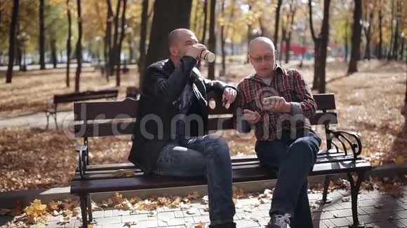 一位年迈父亲的成年儿子在秋公园的长凳上教智能手机视频的预览图