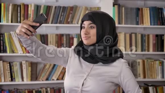 年轻的穆斯林美女戴着头巾自拍然后在图书馆看电话和看照片微笑着视频的预览图