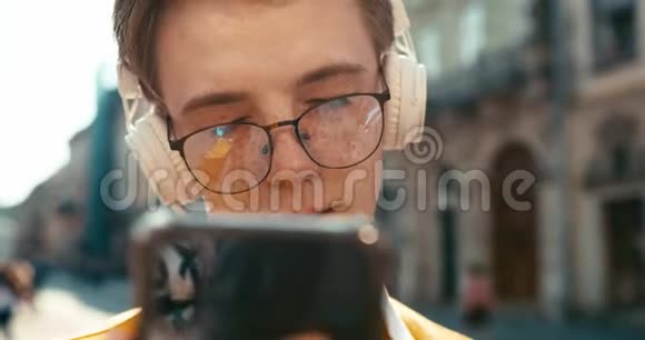 前面的特写镜头可以看到迷人的微笑的嬉皮士戴着眼镜拿着智能手机享受着音乐视频的预览图
