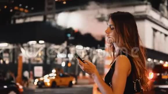 年轻漂亮的女人晚上站在纽约拥挤的交通街道上使用智能手机视频的预览图