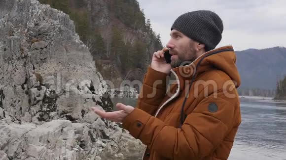 年轻的帅哥在寒冷的天气里站在河边胡子男在打电话慢动作视频的预览图