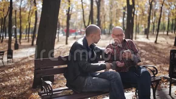 一位年迈的父亲和他成年的儿子坐在公园的长凳上喝咖啡和说话年长父亲的成年儿子视频的预览图