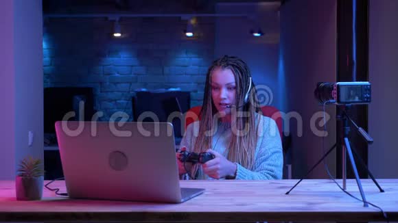在笔记本电脑和电脑上玩电子游戏的耳机里戴着可怕的耳机的年轻有魅力的女性博主特写镜头视频的预览图