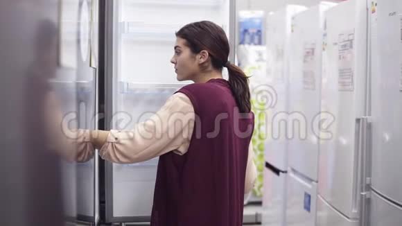 年轻的黑发女孩在五金店寻找完美的冰箱她检查了白色冰箱里的看上去视频的预览图