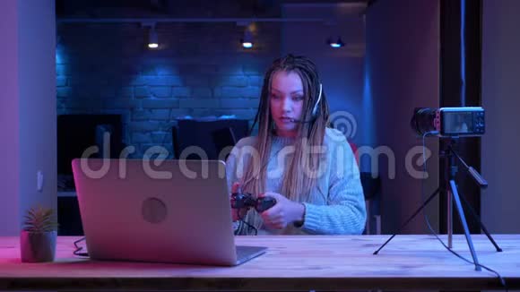 在笔记本电脑和电脑上玩电子游戏的耳机里戴着可怕的耳机的年轻有魅力的女性博主特写镜头视频的预览图