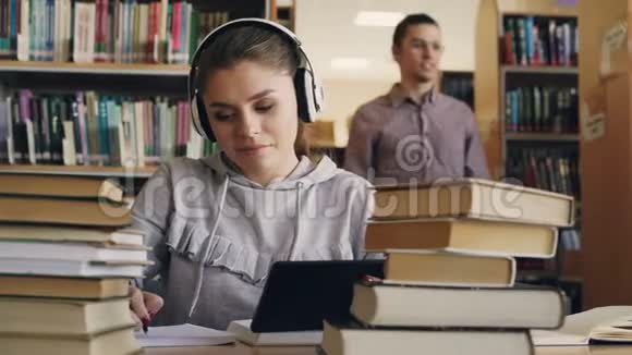 戴着白色耳机的积极微笑的年轻女子坐在大学图书馆的桌子旁手里拿着数码平板电脑和视频的预览图