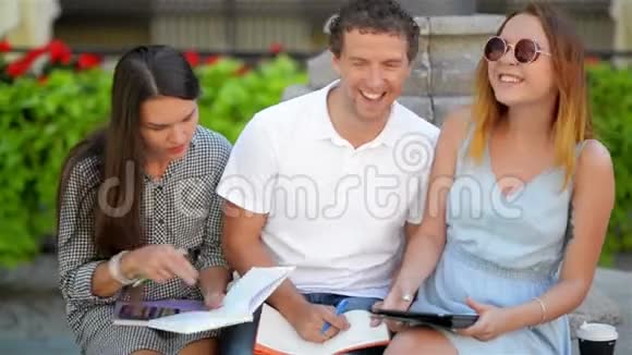 两名女学生与一个男孩一起学习户外坐在长凳上的城市公园电子学习概念视频的预览图