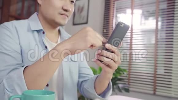 年轻的亚洲男人一边使用智能手机一边躺在客厅的桌子上视频的预览图