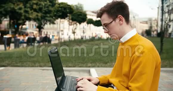 在户外笔记本电脑上工作的戴着眼镜的年轻漂亮金发男人的侧面肖像4K录像视频的预览图