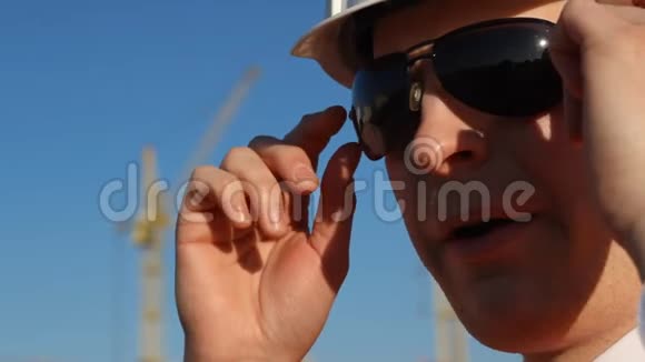 一个戴着黑色眼镜戴着白色头盔嚼口香糖在建筑背景上固定眼镜的人起重机视频的预览图