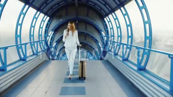 穿着白色西装的时尚商务女性穿过隧道或者从玻璃上过渡带轮子的幸运包视频的预览图