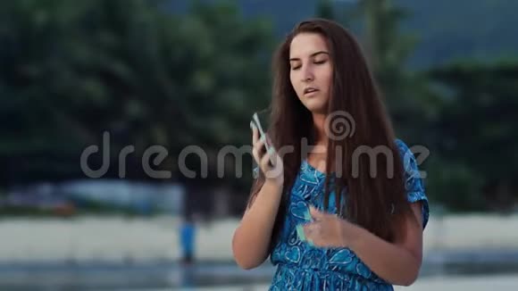 快乐的年轻女孩一头长发穿着一件蓝色的衣服在阳光明媚的海岸棕榈树和打电话视频的预览图