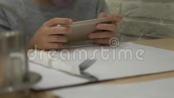 一个孩子正在桌子上玩手机在咖啡馆里等待订单男孩坐在桌子旁抱着视频的预览图