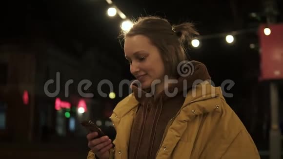 一个漂亮的女孩在一条黑暗的街道中间灯光下看着电话微笑着黄色外套侧视图视频的预览图