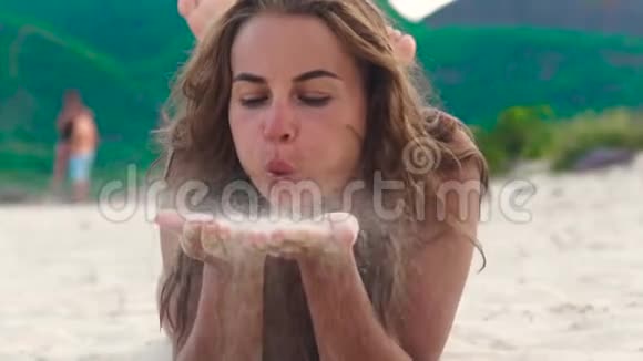 年轻女子在夏天的海滩上用手吹沙面对美丽的女人用手吹着青山上的沙视频的预览图