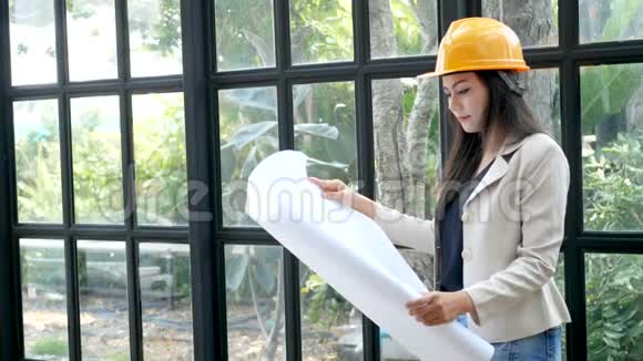 年轻的亚洲女建筑工程师戴着黄色的安全帽手里拿着建筑师的蓝图正在阅读视频的预览图