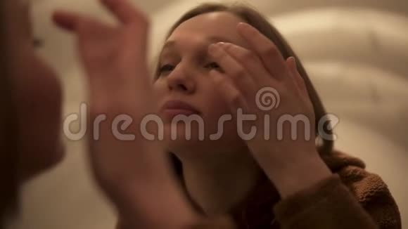 年轻漂亮的浅棕色头发的女孩正在眼睑上涂皱纹霜照镜子手持录像视频的预览图