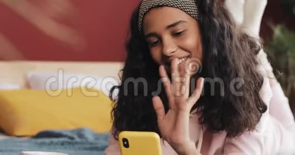 快乐的非裔美国女孩躺在床上她和朋友们在视频中愉快地聊着她的黄色电话视频的预览图