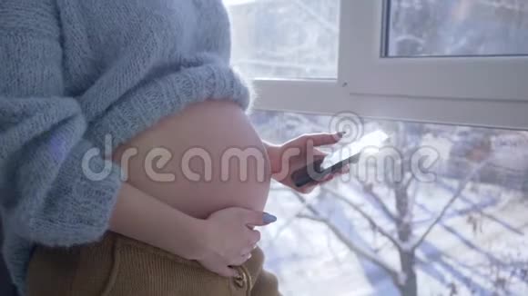孕妇在家中站在一扇大窗户旁用手机抚摸自己赤裸的肚子视频的预览图