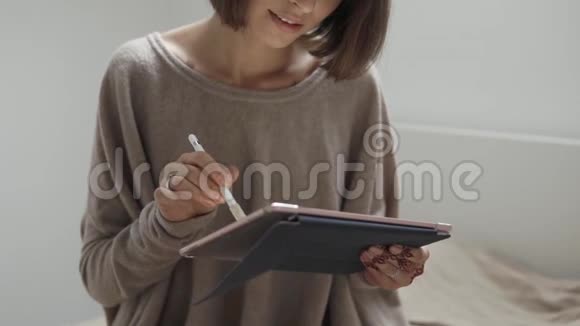 年轻的微笑女性白天在卧室里使用手写笔和平板电脑视频的预览图