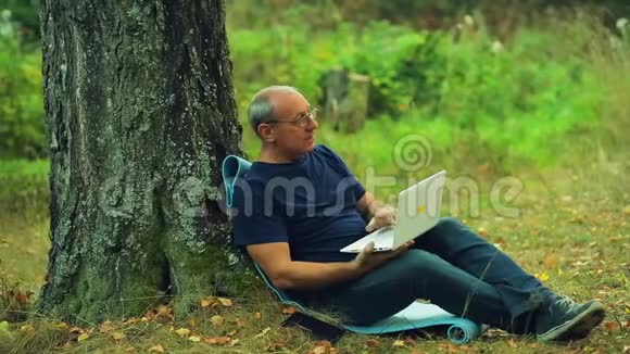 戴眼镜的人坐在公园的一棵树下用笔记本电脑工作视频的预览图