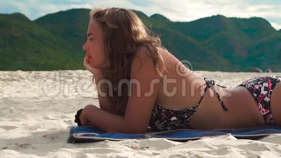 年轻女子在青山背景下的夏季海滩上日光浴穿比基尼的美女在沙滩上晒黑视频的预览图