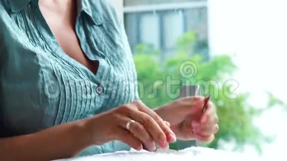 年轻的女人坐在窗户附近在家里做指甲女性在家中使用指甲工具切割角质层视频的预览图