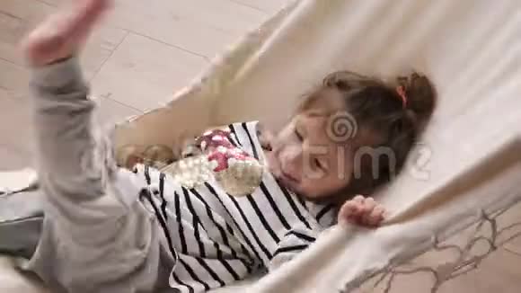 快乐的小女孩在室内玩躺在吊床上挥舞着她的腿微笑的孩子视频的预览图