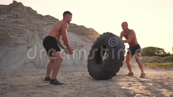 两个肌肉发达的开胸运动员在海滩上以主动的方式训练做推UPS和推一个巨大的轮子对抗一个视频的预览图