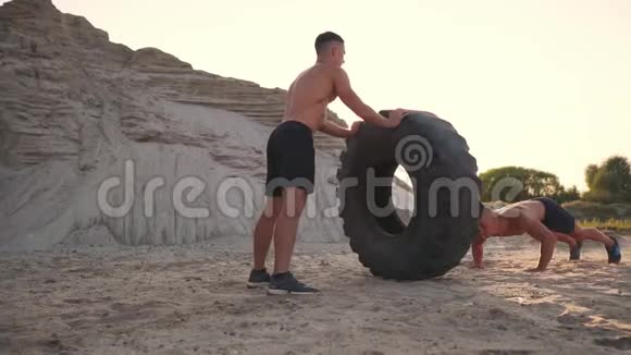 两个肌肉发达的开胸运动员在海滩上以主动的方式训练做推UPS和推一个巨大的轮子对抗一个视频的预览图