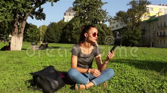 美丽的年轻女孩坐在公园的草地上在电话里听她最喜欢的曲调视频的预览图