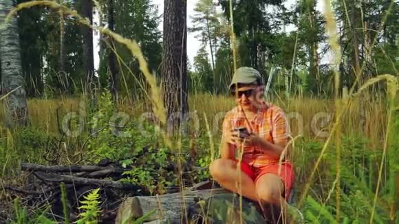 一位年轻的女游客戴着太阳镜和棒球帽坐在一棵倒下的树上在电话里交流视频的预览图