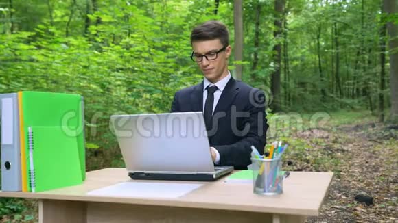 穿西装的严肃商人在森林清新的空气里的办公桌上用笔记本电脑工作视频的预览图