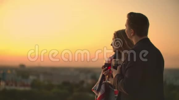 一对年轻夫妇站在屋顶上喝着杯子里的酒站在毯子里欣赏着美丽的日落视频的预览图
