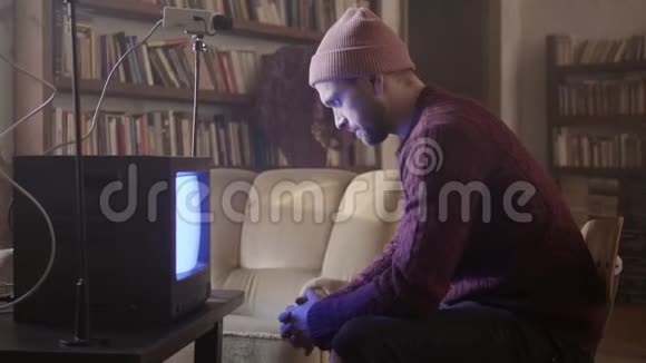 穿毛衣的年轻人坐在家具齐全的房间里的复古电视前的凳子上视频的预览图