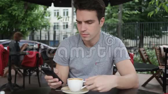 男人一边喝咖啡一边用智能手机坐在咖啡厅露台上视频的预览图