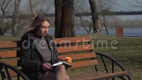 漂亮女孩在公园的长凳上吃苹果在笔记本上做笔记视频的预览图