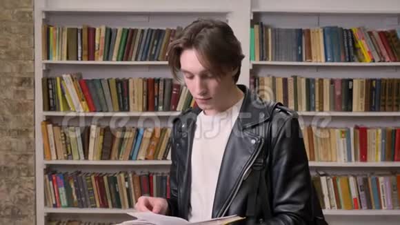 穿黑色皮夹克的年轻男学生站在图书馆看书书架背景视频的预览图