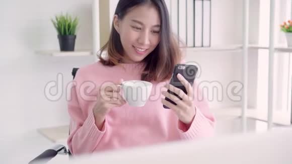 亚洲女性穿着智能休闲服使用智能手机一边喝着温暖的咖啡一边坐在创意办公室的桌子上视频的预览图