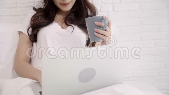 美丽的亚洲女人在卧室里躺在床上玩电脑或笔记本电脑快乐的女性一边喝咖啡一边买视频的预览图