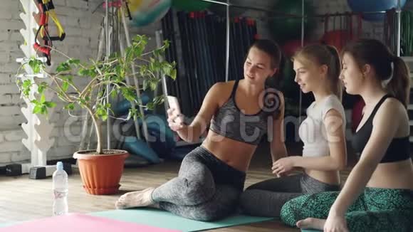 漂亮的女孩在瑜伽工作室里用智能手机自拍年轻的女人在聊天摆姿势笑她们在视频的预览图
