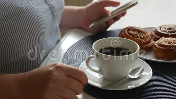 女人坐在桌子旁喝着咖啡夹着肉桂面包端着咖啡看着智能手机视频的预览图