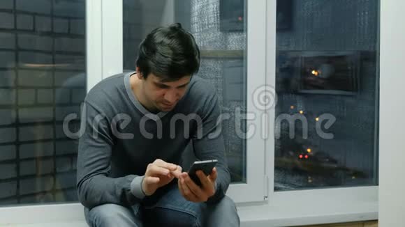 年轻人坐在窗台上浏览他的手机视频的预览图