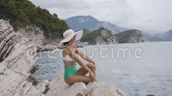 戴着帽子和泳衣的年轻女孩坐在岩石上看着蓝色的大海迷人的女人在海边放松夏季视频的预览图