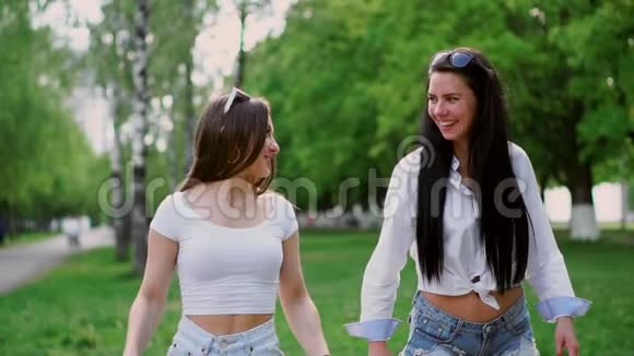 阳光明媚的夏日两个笑眯眯的漂亮女孩骑在陀螺滑板车公园里视频的预览图