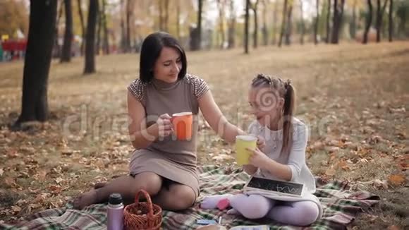 一对可爱的母女正在公园里坐在格子布上喝茶小女孩在黑板上画着蜂视频的预览图