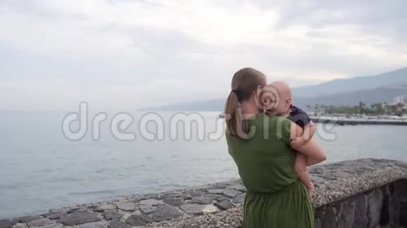 一位年轻的妈妈带着一个婴儿在她的手上射击看着海洋和海浪的惊人景色显示孩子视频的预览图