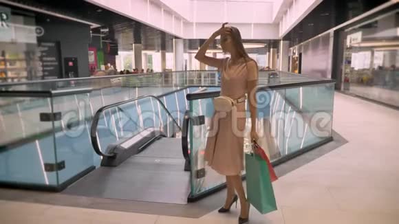 年轻漂亮的女孩站在商场里背着袋子看着摄像机摸着头发购物理念时尚视频的预览图